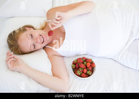 Pregnant woman lying in bed with bowl of strawberries smiling Stock Photo