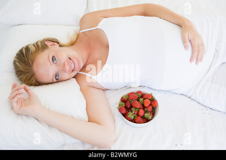 Pregnant woman lying in bed with bowl of strawberries smiling Stock Photo