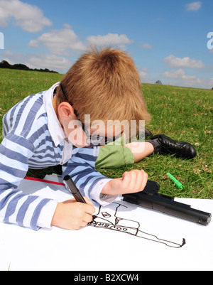 Vertical image of a small boy drawing a picture of his toy gun in the park on a summers day in Kent England Stock Photo