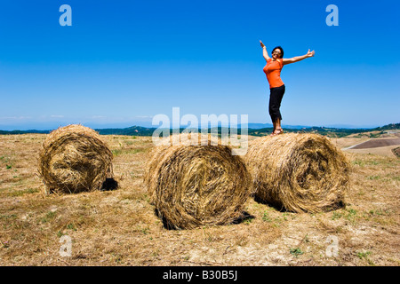 Middle aged woman depicting freedom by standing on rolls of hay in farm field during the summer time in Tuscany Italy Stock Photo