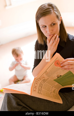 mother looking at worrying financial news in the paper with baby in the background Stock Photo