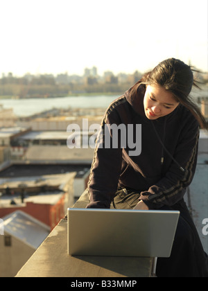 A young Asian woman types on a laptop while sitting on a rooftop. Stock Photo