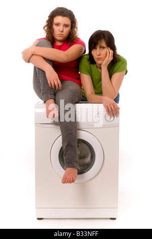 Two casually dressed caucasian females, one leaning and one sitting on a washing machine looking glum. Stock Photo