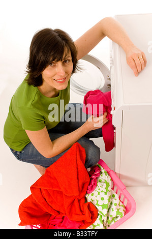 Young caucasian woman wearing a green t-shirt and jeans loading domestic washing machine with brightly coloured clothes from laundry basket. Stock Photo