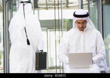 Businessman outdoors using laptop with businessman walking in background (selective focus) Stock Photo