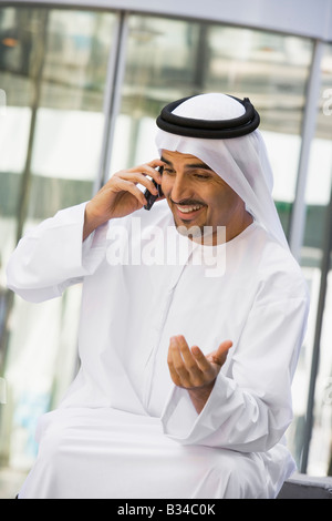 Businessman sitting outdoors by building using cellular phone and smiling (selective focus) Stock Photo