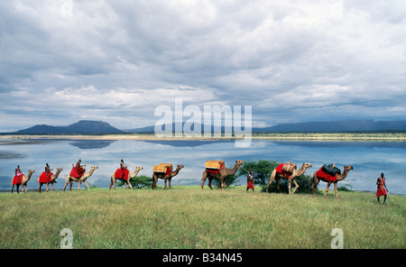 Kenya, Rift Valley Province, Magadi. Maasai men lead a camel caravan laden with equipment for a 'fly camp' (a small temporary camp) past Lake Magadi. Clouds hang low over the Nguruman Escarpment (a western wall of the Great Rift Valley) in the distance. Stock Photo