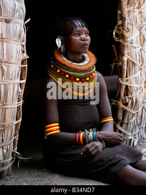 Kenya, Northern Frontier District, Loyangalani. A Turkana woman, typically wearing many layers of bead necklaces and a series of hooped earrings with an pair of leaf-shaped earrrings at the front, sits in the entrance to her hut. Stock Photo