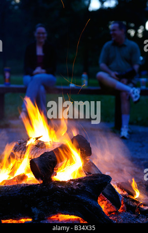 Man and a woman sitting on a bench by a flaming campfire at dusk talking and laughing Stock Photo