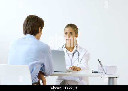 Female doctor sitting across from male patient at desk, using laptop computer Stock Photo