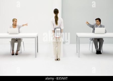 Office workers sitting at desks, both pointing at woman holding document behind her back Stock Photo