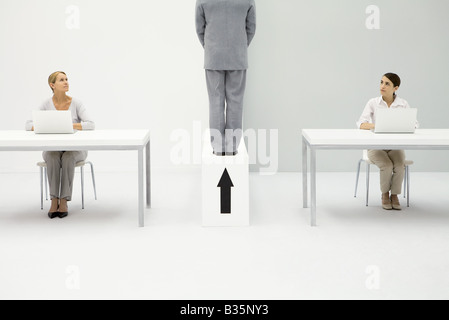 Two women sitting at desks, both looking at man standing above upward pointing arrow Stock Photo