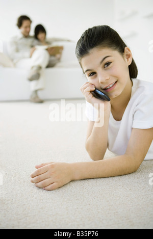 Girl using cell phone, lying on stomach, her family sitting on sofa in background Stock Photo