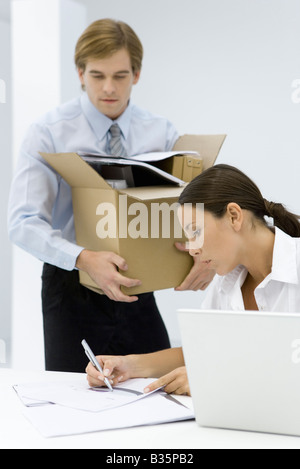 Woman writing on document at desk, man carrying cardboard box full of office supplies Stock Photo