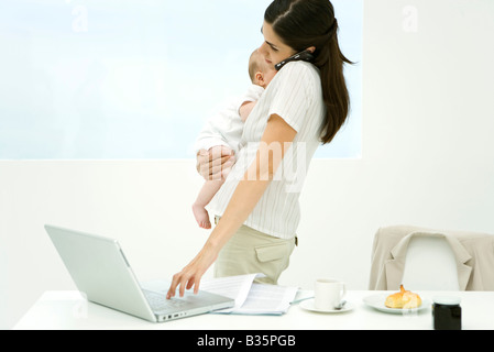 Professional woman standing beside breakfast table, holding baby, using cell phone and laptop computer Stock Photo