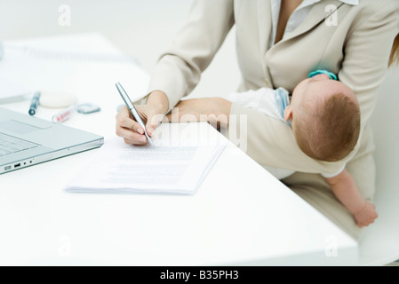 Professional woman signing document at desk, holding sleeping infant, cropped view Stock Photo