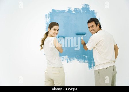 Couple painting wall blue using rollers, both looking over their shoulders and smiling at camera Stock Photo