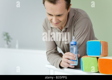 Father leaning against crib, holding baby bottle, looking down Stock Photo