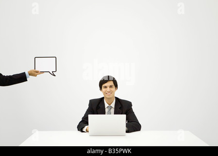 Young businessman sitting with laptop, smiling at camera, colleague holding up blank word bubble Stock Photo