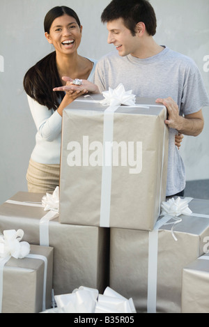 Couple standing behind stack of large gift boxes, man holding tiny present, woman laughing