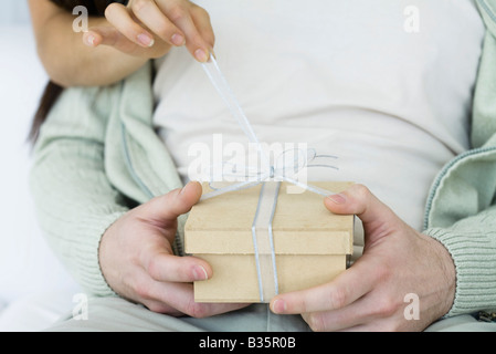 Man holding gift, woman's arm reaching around to untie the ribbon, cropped view Stock Photo
