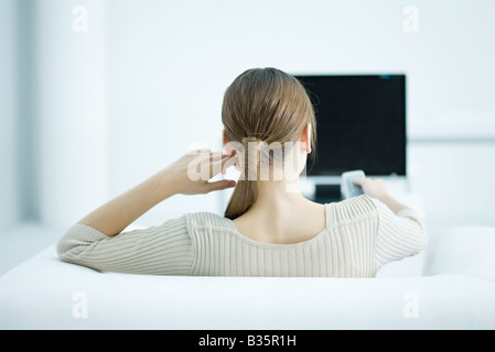 Young woman sitting in armchair watching TV, holding remote, rear view Stock Photo