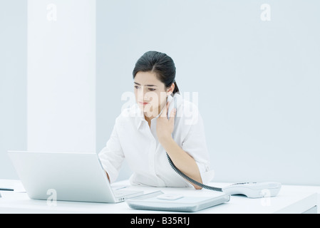 Young professional woman sitting at desk, using laptop, holding landline phone to her chest Stock Photo