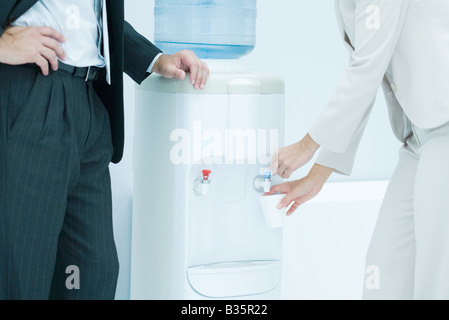 Professional woman filling disposable cup with water from water cooler, male colleague standing nearby, cropped view Stock Photo