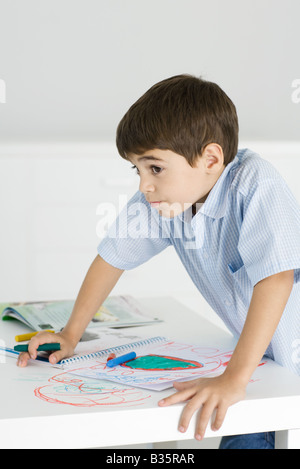 Boy leaning on table, holding crayon, colorful drawing on paper and the table Stock Photo