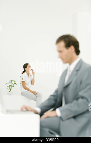 Young woman sitting with cell phone, professional man using laptop computer in foreground Stock Photo