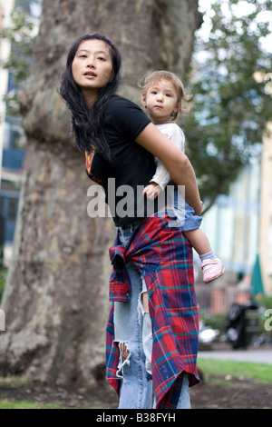 young Korean Asian woman casually dressed carrying mixed race one year old baby on back in London park Stock Photo