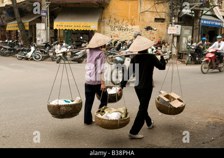 Two women traders with shoulder pole baskets walking up a street in Hanoi Vietnam Stock Photo