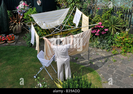 Back garden & lawn of residential property aerial view mature senior woman hanging wet washing on rotary clothes line to dry in sunshine England UK Stock Photo