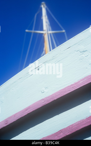 Side of wooden boat showing overlapping timbers painted light grey and pink with shadows and mast against deep blue sky Stock Photo