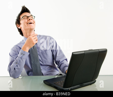 Asian businessman sitting at desk working on laptop computer laughing Stock Photo