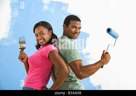 African American couple next to half painted wall as they stand back to back with paint utensils Stock Photo