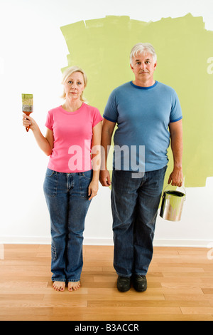 Portrait of unhappy adult couple standing in front of half painted wall with paint supplies American Gothic style Stock Photo