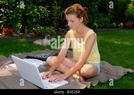 Siamese cat lounging in grass lawn with teenager typing on laptop in back yard shade of a tree beside a garden Stock Photo