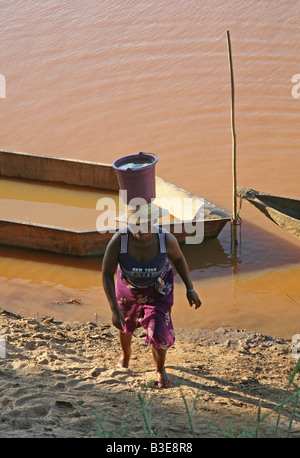 woman is carrying a bucket on her head up a bank slope Frau trägt Wassereimer auf dem Kopf eine Uferböschung hinauf Stock Photo