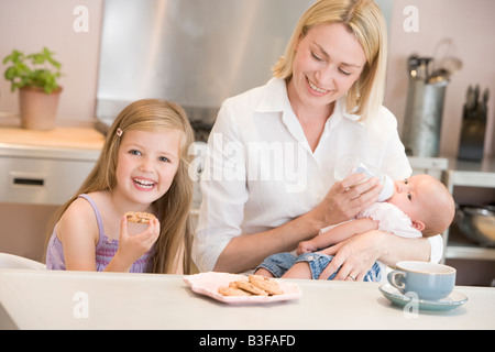 Mother feeding baby in kitchen with daughter eating cookies and smiling Stock Photo