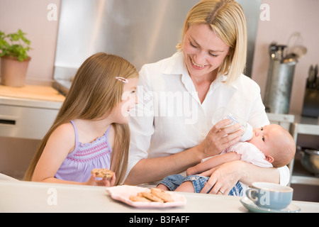 Mother feeding baby in kitchen with daughter eating cookies and smiling Stock Photo