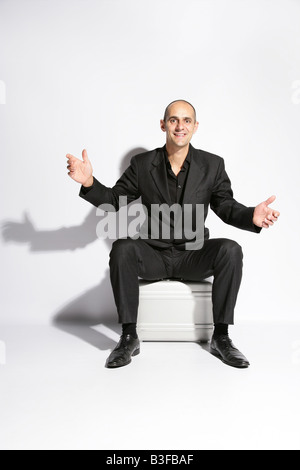 Man in a busines suit sat on a brief case shrugging his shoulders Stock Photo