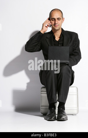 Man in a busines suit sat on a brief case lap top on knee speaking on the telephone Stock Photo