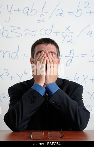 Tired Business Man Rubbing His Eyes Whilst Sitting at His Desk in Front of a Whiteboard Full of Calculations Stock Photo