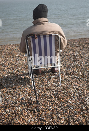 Elderly man from behind wearing coat and hat sitting in chair on shingle beach looking out to sea Stock Photo