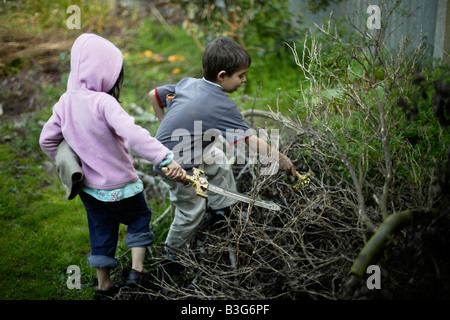 Boy aged six and sister five flush out imaginary foes from pile of tree cuttings with plastic toy swords Stock Photo