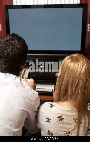 young late teenage couple sitting together playing a video game on a large screen plasma tv in a living room Stock Photo