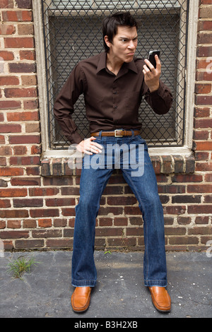 Confused young Asian man next to brick wall and window looking at cell phone Stock Photo