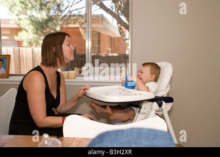 mom making baby laugh in high chair during snack feeding, making funny faces, baby smiles and giggles back at mother Stock Photo