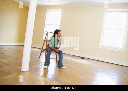 Man sitting on ladder in empty space holding paper thinking Stock Photo